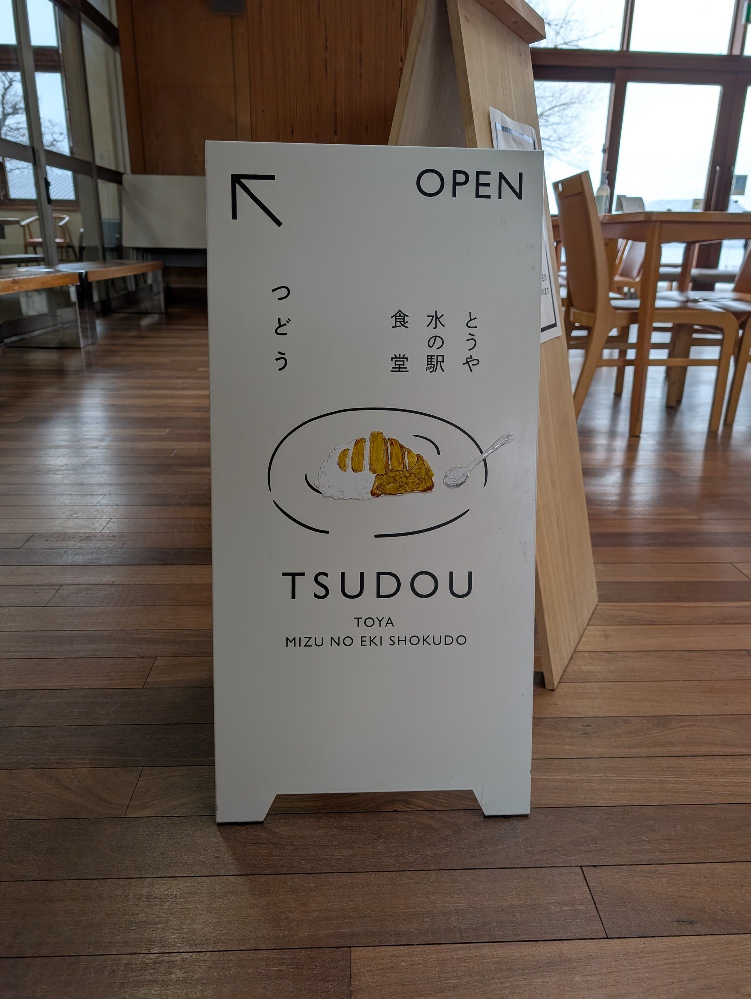 TSUDOUの看板。白い立て看板にカレーのイラストと「つどう とうや水の駅食堂 TSUDOU TOYA MIZU NO EKI SHOKUDO」の文字