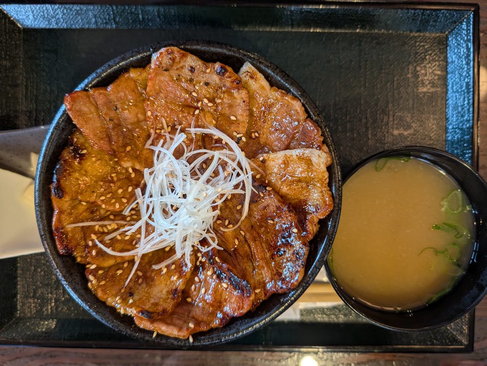 ランチセット1の炭火焼た丼タレ。黒い鉄鍋に艶やかなタレの豚バラ肩ロース、千切りネギとゴマが散らされている